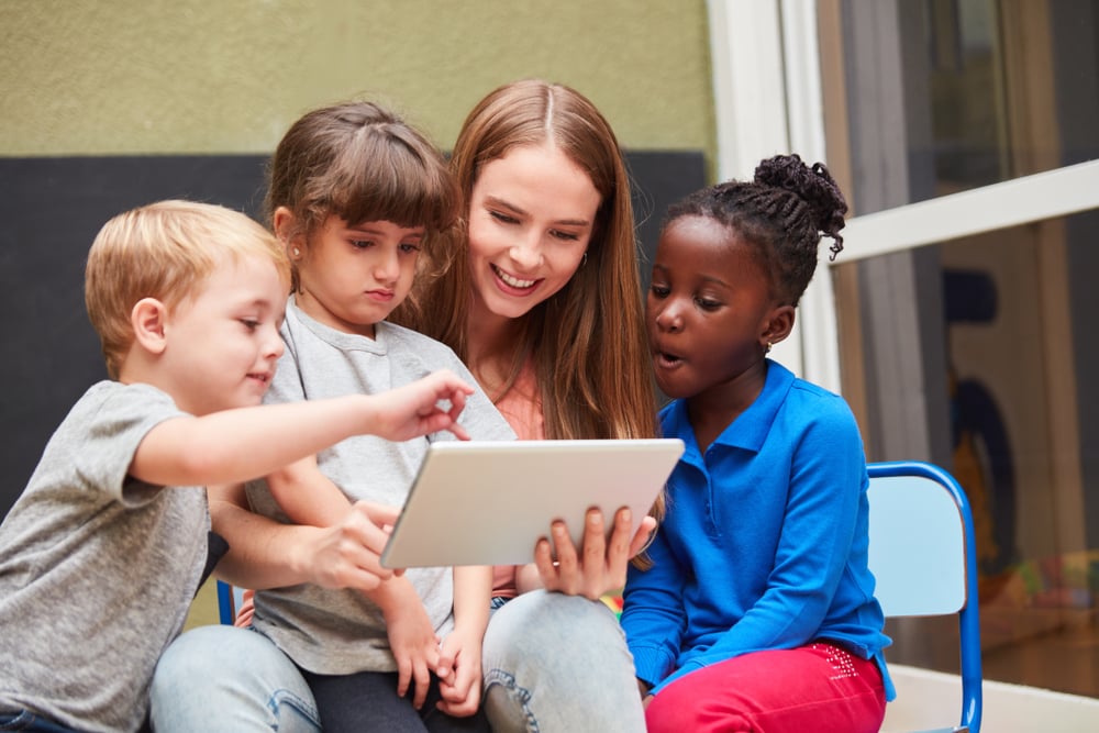 Profissional de Pedagogia sentada junto a crianças interagindo com um tablet